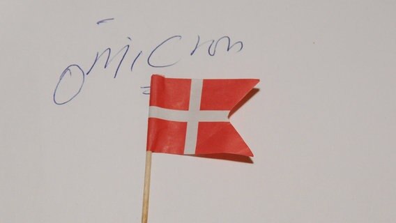 Ein Zahnstocher mit einer Dänemarkflagge liegt auf einem Blatt Papier wo "omicron" drauf steht. © imago images Foto: Francis Joseph Dean