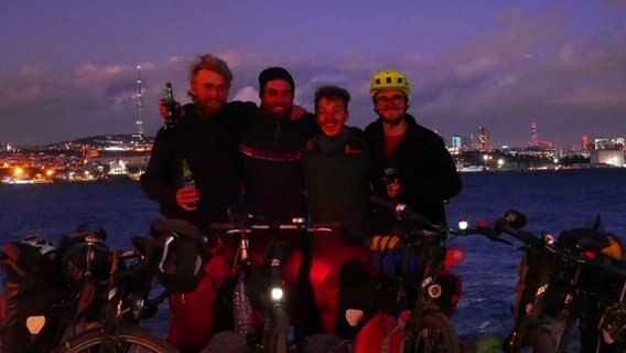 Vier Radfahrer stehen bei Nacht mit ihren Fahrrädern und Getränken in der Hand am Wasser. Im Hintergrund ist eine Skyline zu sehen.  Foto: Projekt Open Eyes