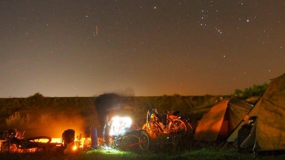 Es ist ein Sternenhimmel zu sehen. Ein Mann steht bei Fahrrädern und zwei Zelten.  Foto: Projekt Open Eyes