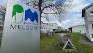 Eine Tafel mit dem Logo der Papierfrabrik Meldorf sowie eine Papierrolle auf einem speziellen Stönder stehen auf dem Rasen auf dem Gelände der Papierfabrik in Tornesch. © NDR Foto: Christoph Deuschle