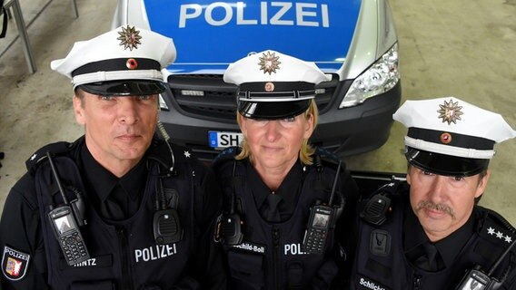 Drei Polizisten präsentieren ihre neuen weißen Polizeihüte und stichfesten Westen. Im Hintergrund steht einen Polizeiwagen © dpa-Bildfunk Foto: Carsten Rehder