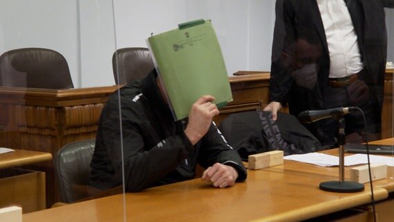 Ein Angeklagter sitzt vor Gericht und verdeckt sein Gesicht mit einem Aktenordner.  