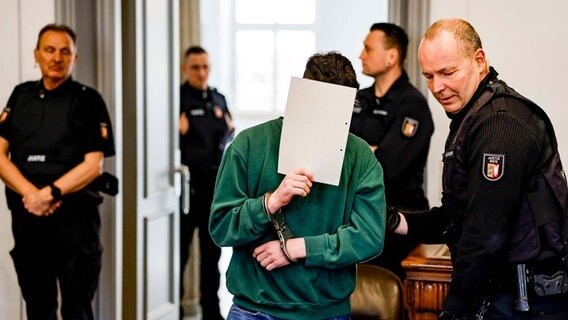 Ein junger Mann verdeckt in einem Gerichtssaal zwischen Beamten sein Gesicht mit einem Papier. © dpa Foto: Axel Heimken