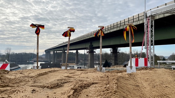 An der Rader Hochbrücke stecken auf einer Baustellen vier Spaten nebeneinander im Sand. © NDR Foto: Christopher Gaube