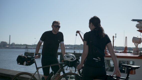Ein Radfahrer und eine Radfahrerin stehen vor einem Hafenbecken. © NDR Foto: Dominik Dührsen