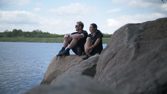 Ein Mann und eine Frau sitzen auf großen Steinen am Wasser. © NDR Foto: Dominik Dührsen