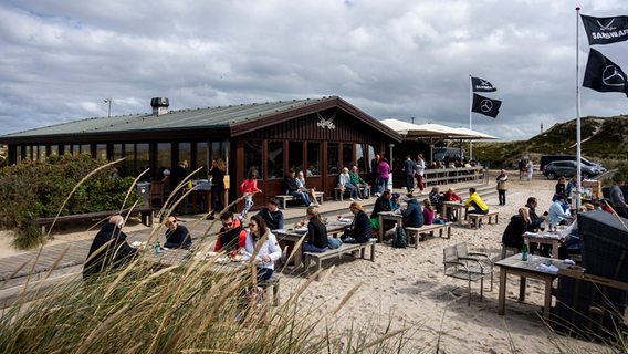 Gäste sitzen an Tischen draußen auf dem Gelände der Strandbar Sansibar auf der Nordseeinsel Sylt (Schleswig-Holstein). © picture alliance Foto: Jürgen Schwenkenbecher