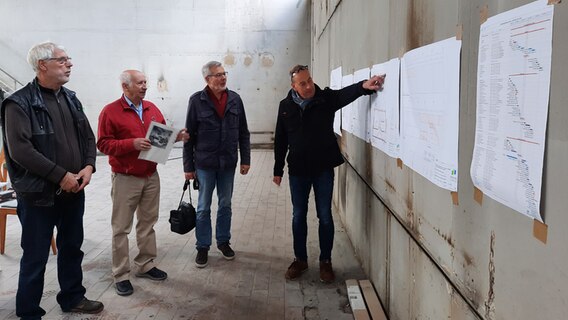 Vier Männer stehen in einem Großraum eines Gebäudes in Nortorf an Bauzeichnungen, die an einer Wand. © NDR Foto: Lina Bande