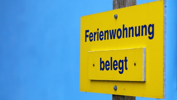 Vor einer Ferienhaus steht ein Hinweisschild mit dem Schriftzug "Ferienwohnung belegt". © dpa Foto: Arno Burgi