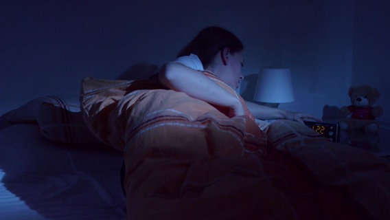 Eine Frau liegt im Bett und schaut auf ihren Wecker im Dunkeln. © NDR 