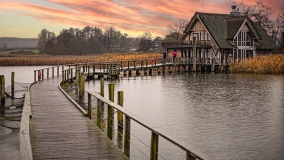 Ein Holzsteg auf dem Hemmelsdorfer See fürht zu einem Haus, im Hintergrund färbt sich der Himmel langsam rosa. © Denis Hoppe Foto: Denis Hoppe