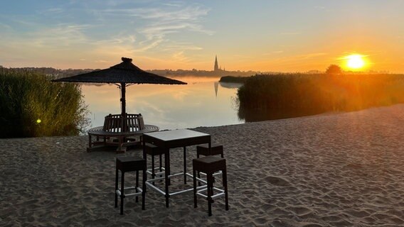 Barhocker und ein Tisch stehen im Sand, am Horizont geht die Sonne auf. © Benjamin Meißner Foto: Benjamin Meißner