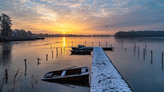 Sonnenaufgang über einem gefrorenen See. Boote stecken neben dem Steg im Eis fest. © Ralf Denguth Foto: Ralf Denguth