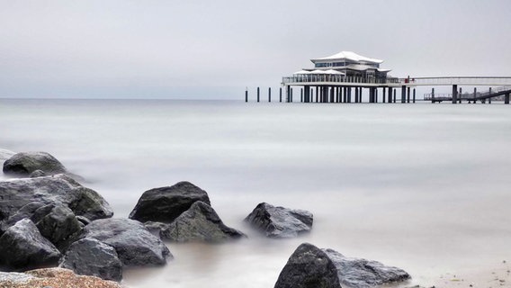 Hinter dunklen Steinen sieht man das Meer, einen bewölkten Himmel und ein auf Pfählen gebautes Gebäude. © Karin Mädlow Foto: Karin Mädlow