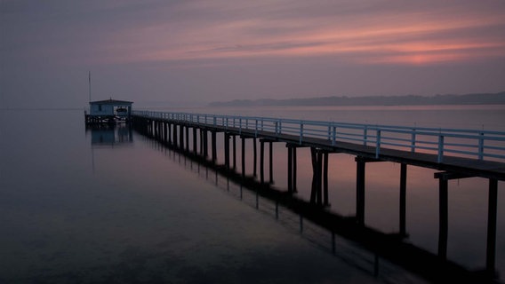 Das von der Sonne gefärbte Wasser der Förde und ein Steg, der zu einem Holzhaus mit Boot führt. © Piotr Mankowski Foto: Piotr Mankowski