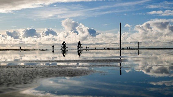 Zwei Fahrradfahrer fahren von links nach rechts durchs Bild, auch einige Fußgänger sind unterwegs. Es ist blauer Himmel mit strahlend weißen Wolken, welche mit den Menschen zusammen im Wasser gespiegelt werden. © Wenke Stahlbock Foto: Wenke Stahlbock