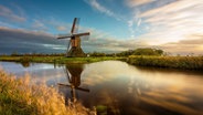 Eine Mühle steht hinter einem stillen Gewässer, welches das blau-gelbe Farbspiel des Himmels spiegelt. Um das Wasser herum sind sattgrüne Felder und Pflanzen. © Björn Behrendsen Foto: Björn Behrendsen