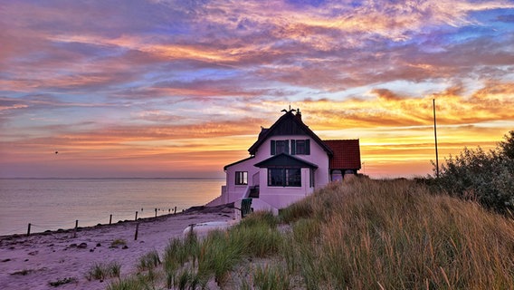 Ein scheinbar einsames Haus am Ufer. Hintergrund das Wasser, der Himmel in bunten Farben des Sonnenuntergangs. Vorne im Bild wehen grüne Gräser im Wind. © Michael Buske Foto: Michael Buske