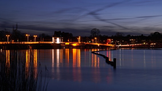 Die Kappelner Brücke mit ihrer Beleuchtung spiegelt sich im Dunkeln im Wasser der Schlei. Im Vordergrund ist der Heringszaun zu sehen. © Heike Greggersen Foto: Heike Greggersen