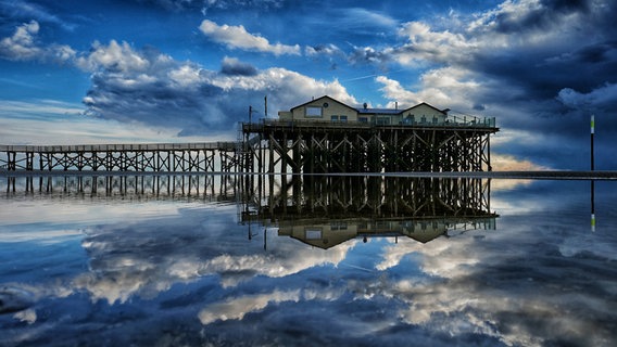 Ein Stelzenhaus mit Brücke spiegelt sich im Wasser der Nordsee auf St. Peter Ording. © Achim Otto Foto: Achim Otto