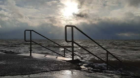 Zusehen ist eine Treppe die am Husumer Dockkoog ins aufgewühlte Meer führt. Der Himmel ist bedeckt mit Wolken. In der Mitte bricht die Wolkendecke und die Sonne kommt durch.  Foto: Ulf Harring-Petersen