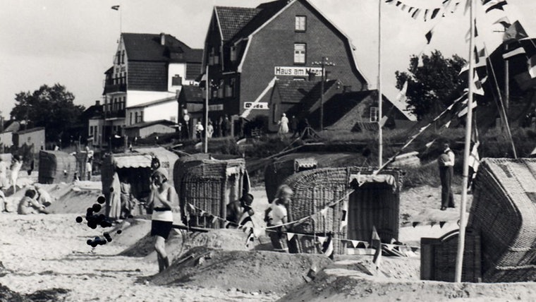 Das historische Bild zeigt den Strand von Schönberg voller Badegäste mit kleinen Wällen um die Strandkörbe herum, Wimpelchen wehen dazuwischengespannt im Wind, Kinder spielen im Sand. © Projekt Spurensuche