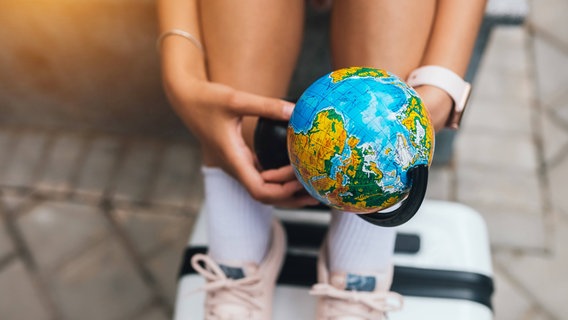 Eine junge Frau hält einen Globus in der Hand und hat ihre Füße auf einem Koffer abgestellt. © imago images / Zoonar 