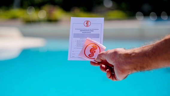 Eine Hand hält ein Seepferdchenabzeichen vor ein Schwimmbecken © Imago Images Foto: Patrick Scheiber