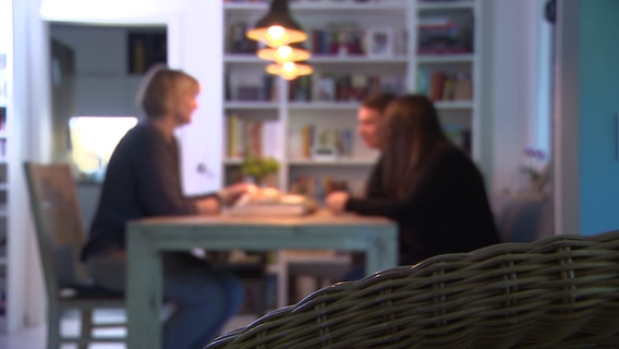 Zwei Frauen und ein Mann sitzen an einem Tisch in einem Wohnzimmer. © NDR 