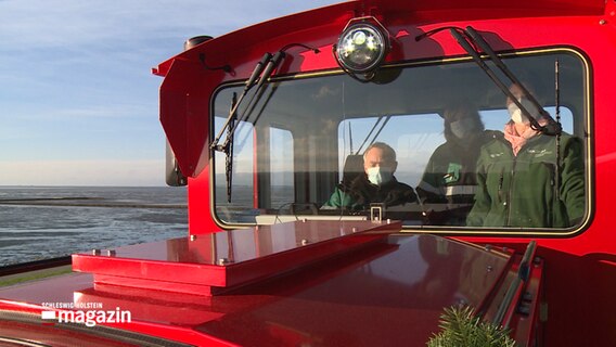 Ein Mann und zwei Frauen befinden sich in der Führerkabine einer roten Lokomotive die über einen Deich vor der Küste fährt. © NDR 