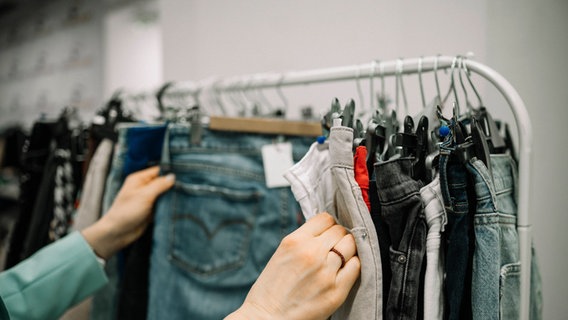 Eine Frau guckt durch verschiedene Jeanshosen an einem Kleiderständer. © imago images Foto: Westend61