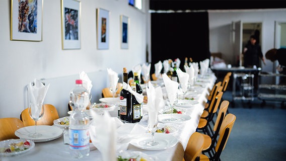 In einem Saal steht eine lange Tafel, an der viele Stühle stehen und die mit Geschirr und Flaschen gedeckt ist. © NDR Foto: Lisa Pandelaki