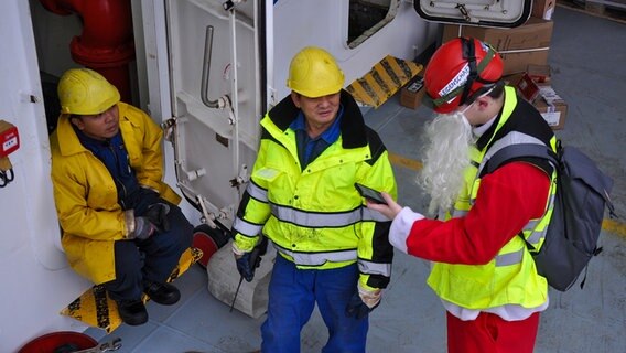 Ein als Weihnachtsmann gekleideter Mann spricht mit zwei Seeleuten am Bord eines Schiffes in Brunsbüttel. © NDR Foto: Pauline Reinhard