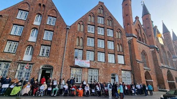 Bewohner sitzen in Rollstühlen vor dem Heiligen-Geist-Hospital, eingepackt in dicke Decken, halten Plakate hoch. Etwa 80 Senioren und Angehörige demonstrieren. © NDR Foto: Julian Marxen