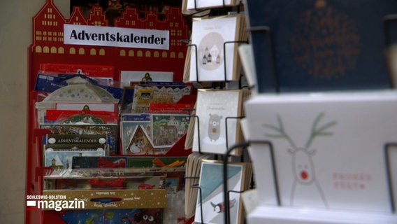 In einem Verkaufsständer sind Postkarten mit Weihnachtsmotiven, im Hintergrund werden Adventskalender angeboten. © NDR Foto: NDR Screenshots