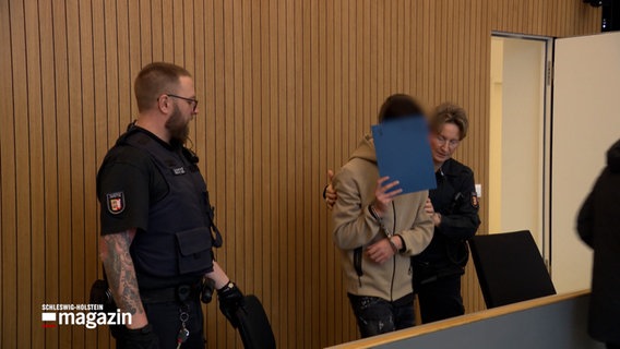 Ein Angeklagter wird in einem Gerichtssaal gebracht, er verdeckt sein Gesicht mit einer blauen Mappe. © NDR Foto: NDR Screenshot