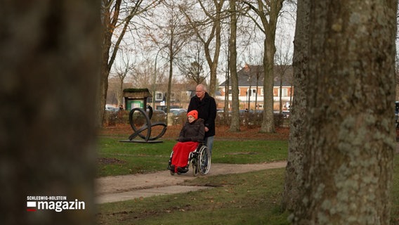 Ein Mann schiebt eine Frau im Rollstuhl durch einen Park. © NDR Foto: NDR Screenshot