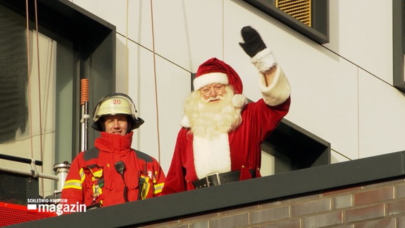 Der Weihnachtsmann winkt von einem Dach, neben ihm steht ein Feuerwehrmann. © NDR Foto: NDR Screenshot