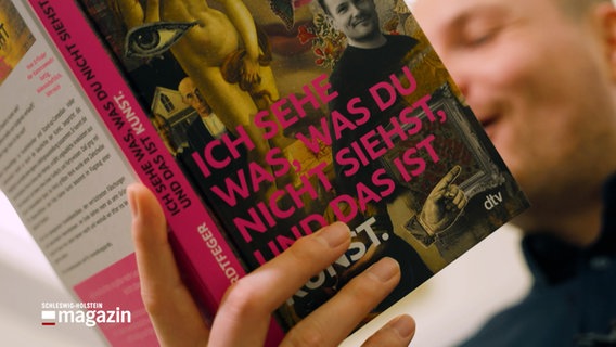 Ein Mann liest in einem Buch mit dem Titel: "Ich sehe was, was du nicht siehst, und das ist Kunst." © NDR Foto: NDR Screenshot