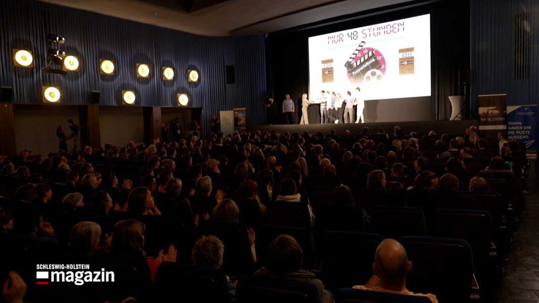 Mehrere Personen gehen von einer Bühne, auf der Bühne wird über einen Projektor das Logo des Filmwettbewerbes 