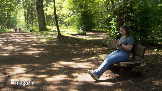 Eine Frau sitzt auf einer Bank in einem Wald und schaut auf ihr Smartphone © NDR Foto: NDR Screenshot