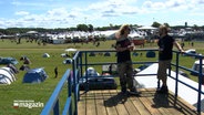 Zwei Personen stehen auf einer Dachterrasse vor dem Campingplatz des Wacken Open Air © NDR Foto: NDR Screenshot