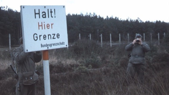 Eine alte Videoaufnahme zeigt ein Schild mit der Aufschrift "Halt! Hier Grenze / Bundesschutz". Ein Grenzschutzsoldat fotografiert währenddessen den Kameramann.  