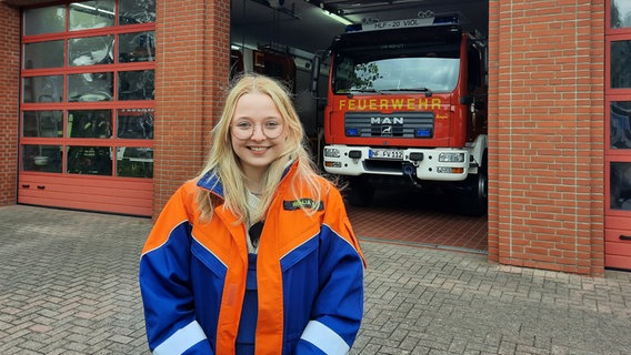 ist Ronja Sievertsen, Ü18-Siegerin bei "Vertell doch mal" steht vor einer Feuerwehrhalle mit einem Feuerwehrfahzeug und Trägt eine Feuerwehrjacke. © NDR Foto: Lina Bande