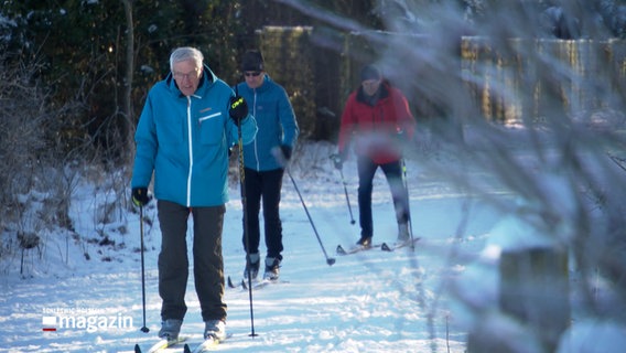Ältere Leute fahren mit Ski über eine Straße. © NDR 