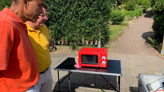 Zwei Männer blicken auf eine Mkrowelle, die durch ein Solar-Panel betrieben wird. © NDR Foto: Lena Haamann