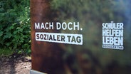 Ein Banner mit dem Schriftzug "Mach doch. Sozialer Tag" steht auf einem Schotter-Boden. © NDR Foto: Johannes Tran
