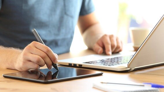 Ein Mann malt auf ein Tablet, während er parallel einen Laptop bedient. © imago images / Panthermedia Foto: Antonio Guillem