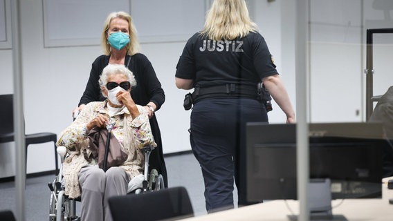 Eine Frau im Rollstuhl wird von einer anderen in den Gerichtssaal gefahren. Eine Mitarbeiterin der Justiz steht daneben. © Christian Charisius/dpa Foto: Christian Charisius/dpa