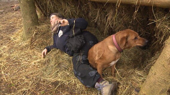 Eine Frau liegt in einem Unterschlupf mit Stroh mit ihrem Hund bei einem Survival-Training in einem Wald. © NDR 